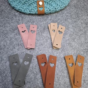 Könnte beinhalten: Eine Sammlung von Lederetiketten in verschiedenen Farben, darunter Rosa, Beige, Grau, Braun und Hellbraun. Jedes Etikett hat einen herzförmigen Ausschnitt und ein Loch zur Befestigung. Im Hintergrund ist ein türkisfarbenes Häkelteil mit einem braunen Lederetikett zu sehen.