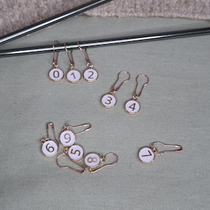 Könnte beinhalten: Zehn goldene Maschenmarkierer mit weißen Emaille-Zahlen von 0 bis 9. Die Markierer haben die Form eines Kreises mit einer kleinen Schlaufe oben.