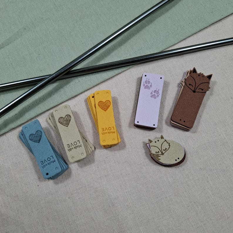 Op de afbeelding: Een verzameling kleine, decoratieve labels en houten versieringen. De labels zijn in verschillende kleuren, waaronder blauw, geel en beige, met hart- en pootafdrukontwerpen. De houten versieringen hebben vossen. De tekst "Made with LOVE" staat op de labels.