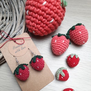Op de afbeelding: Handgemaakte gehaakte aardbeien, waaronder een paar oorbellen. De oorbellen zijn bevestigd aan een label met de tekst "Handmade with love". De aardbeien zijn rood met witte zaadjes en groene blaadjes.