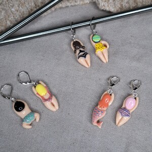 Könnte beinhalten: Sechs Paar Ohrhänger mit Miniaturfiguren in Badebekleidung. Die Ohrringe zeigen verschiedene Designs, darunter eine Meerjungfrau und Figuren in Badeanzügen in verschiedenen Farben. Die Ohrringe sind an silberfarbenen Reifen befestigt.
