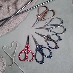 Könnte beinhalten: Eine Sammlung kleiner, dekorativer Scheren in verschiedenen Farben und Designs, darunter Kupfer, Silber und Rot mit weißen Tupfen. Stricknadeln und Garn sind ebenfalls vorhanden, was auf ein Bastelthema hindeutet.
