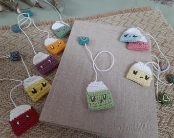 May include: A set of crocheted tea bag bookmarks with different coloured faces and expressions. The bookmarks are attached to a white string and are hanging from a book.