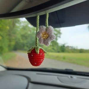 May include: A handmade crochet car charm featuring a red strawberry with white seed details and a white daisy with a yellow center. Both are suspended by green yarn, adding a touch of whimsy to the car's interior.