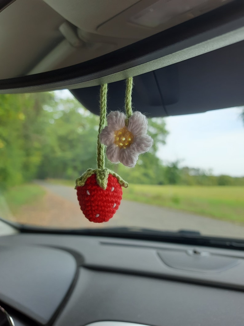 May include: A handmade crochet car charm featuring a red strawberry with white seed details and a white daisy with a yellow center. Both are suspended by green yarn, adding a touch of whimsy to the car's interior.