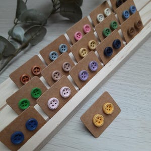 Könnte beinhalten: Sortiment an bunten Knopf-Ohrringen, die auf braunem Karton präsentiert werden. Jedes Paar Ohrringe hat eine andere Farbe und ein anderes Design.
