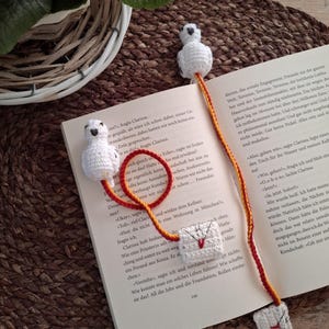 Può includere: Due segnalibri all'uncinetto con gufi bianchi con occhi e becchi neri. I gufi sono attaccati a cordini di filo rosso e giallo. I cordini sono decorati con piccole buste all'uncinetto. I segnalibri sono appoggiati su un libro aperto con testo in tedesco.