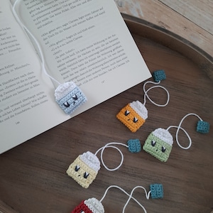 Könnte beinhalten: Ein Set aus sechs gehäkelten Teebeutel-Lesezeichen. Die Teebeutel sind in verschiedenen Farben, darunter Rot, Orange, Gelb, Grün und Blau. Jeder Teebeutel hat ein süßes, gesticktes Gesicht.