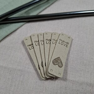 Op de afbeelding: Een stapel lichtbruine rechthoekige labels met afgeronde hoeken. Op elk label staat in zwarte letters "Made with LOVE". Onderaan de labels is een hartvormig ontwerp gedrukt. De labels liggen op een lichtroze ondergrond.