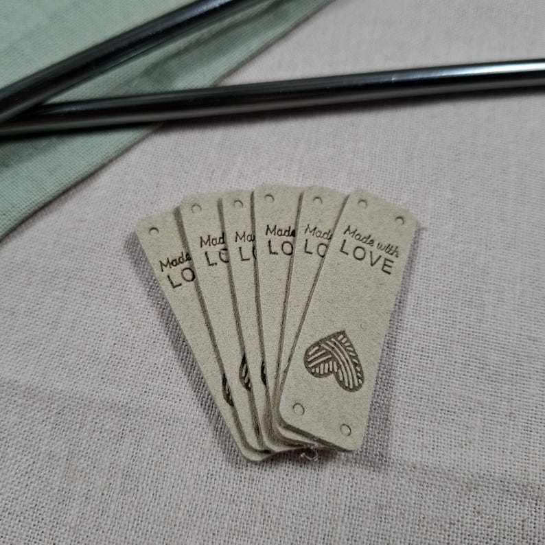 Op de afbeelding: Een stapel lichtbruine rechthoekige labels met afgeronde hoeken. Op elk label staat in zwarte letters "Made with LOVE". Onderaan de labels is een hartvormig ontwerp gedrukt. De labels liggen op een lichtroze ondergrond.