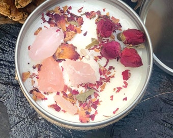 Pode incluir: Uma vela branca com pétalas de rosa, folhas secas e cristais de quartzo rosa em uma lata de prata.