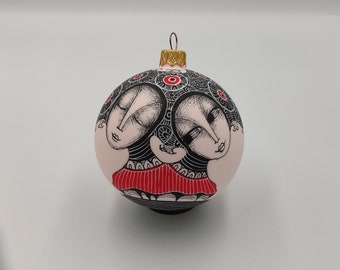 May include: A pink Christmas ornament with a black and white illustration of two women with red accents. The women are facing each other and have their heads tilted.