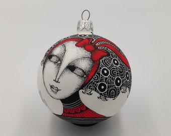 May include: White Christmas ornament with a black and white illustration of a woman with a red bow in her hair. The woman's hair is styled in a complex pattern of circles and swirls.