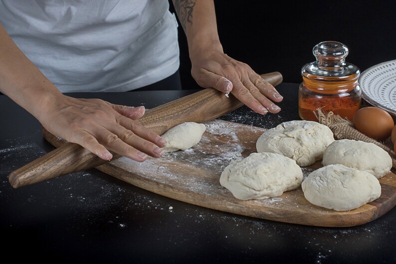 May include: A person rolls out dough on a wooden cutting board with a rolling pin. There are several balls of dough on the board, along with a jar of spices and eggs.