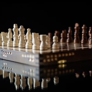 May include: A wooden chessboard with white and brown chess pieces. The board is closed and the pieces are set up for a game. The board is sitting on a black surface.