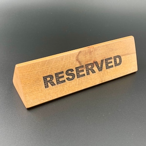 Peut inclure: Une pancarte en bois avec le mot "RESERVED" en lettres noires. La pancarte est de forme triangulaire et a une finition en bois naturel.