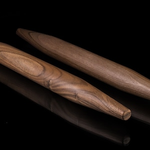 May include: Two wooden rolling pins, one longer than the other, with a dark brown wood grain pattern. The rolling pins are on a black surface.