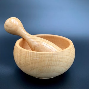 Peut inclure: Un ensemble mortier et pilon en bois clair. Le mortier est en forme de bol et le pilon est arrondi et lisse.