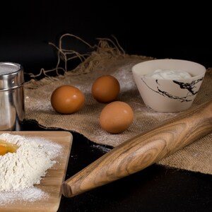 May include: A wooden rolling pin, a bowl of cream, three brown eggs, a metal shaker, and a wooden cutting board with flour and a cracked egg on it. The items are arranged on a burlap surface.