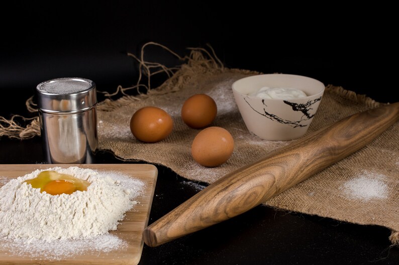 May include: A wooden rolling pin, a bowl of cream, three brown eggs, a metal shaker, and a wooden cutting board with flour and a cracked egg on it. The items are arranged on a burlap surface.