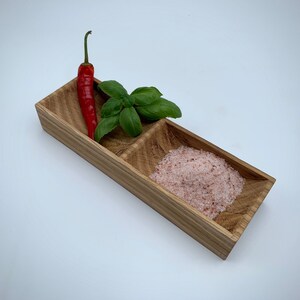May include: A wooden serving tray with two compartments. One compartment contains pink Himalayan salt and the other compartment contains a red chilli pepper and green basil leaves.