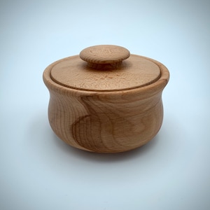 Puede incluir: Una pequeña caja de madera con tapa. La caja está hecha de madera clara y tiene una forma redonda y lisa. La tapa tiene un pequeño pomo en la parte superior.