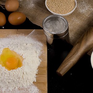 May include: A close-up of a wooden rolling pin, a bowl of white yogurt, a bowl of white quinoa, a metal sifter, and a wooden cutting board with a mound of white flour and a cracked egg yolk.