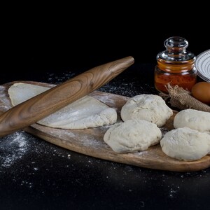May include: A wooden rolling pin sits on a wooden cutting board with five balls of dough and a sprinkle of flour. There is a jar of spices and a white plate with a decorative pattern in the background.