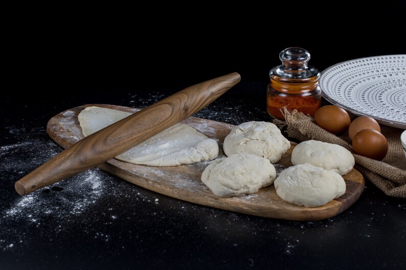 May include: A wooden rolling pin sits on a wooden cutting board with five balls of dough and a sprinkle of flour. There is a jar of spices and a white plate with a decorative pattern in the background.