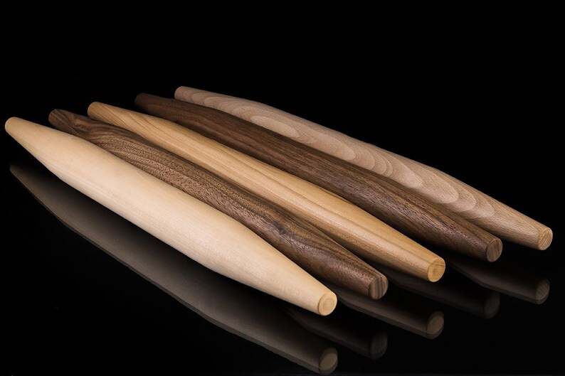 May include: Four wooden rolling pins of varying lengths and diameters, with a light brown, dark brown, and reddish brown wood grain. The rolling pins are arranged in a staggered pattern on a black surface.