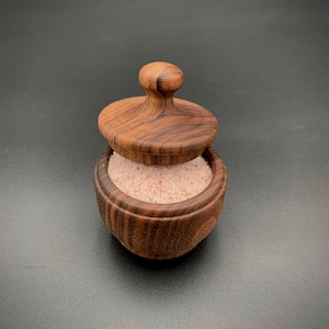 Puede incluir: Un pequeño salero de madera con tapa, lleno de sal rosa del Himalaya. El salero es de madera oscura y tiene forma redondeada. La tapa tiene un pequeño asa en la parte superior. El fondo es negro liso.