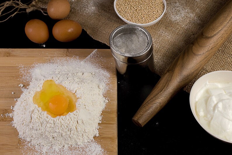 May include: A close-up of a wooden rolling pin, a bowl of white yogurt, a bowl of white quinoa, a metal sifter, and a wooden cutting board with a mound of white flour and a cracked egg yolk.
