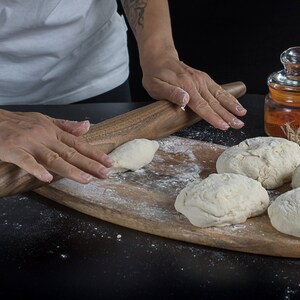 May include: A person rolls out dough on a wooden cutting board with a rolling pin. There are several balls of dough on the board, along with a jar of spices and eggs.