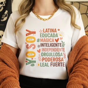 May include: White t-shirt with the words "YO SOY" in orange and green, and a list of positive adjectives in Spanish. The shirt has a crew neck and is paired with a gold chain necklace and a rust-colored cardigan.