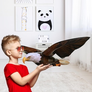 Peut inclure: Un jouet d'aigle marron et blanc aux ailes déployées, tenu par une personne portant un t-shirt rouge et des lunettes de soleil. L'aigle a un bec jaune et se trouve dans une pièce avec des œuvres d'art pour enfants.