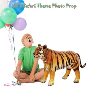 May include: An inflatable tiger prop with orange and black stripes, perfect for a safari-themed photo. The image also includes a child with face paint and a bunch of colorful balloons. Text at the top reads "Kid's Safari Theme Photo Prop."