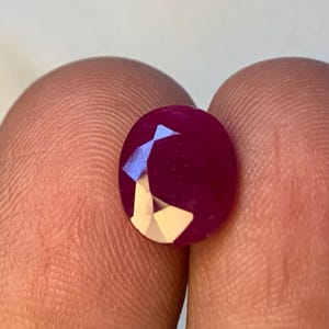 May include: A faceted, oval ruby gemstone. The gem is a deep red colour with a polished surface. The facets create a play of light, highlighting the gem's brilliance. The ruby is held between two fingers.