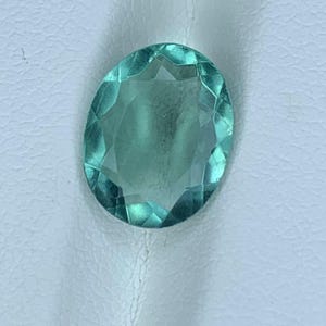 May include: Oval-shaped, faceted gemstone in a light teal color. The gem has a polished surface with multiple facets that catch the light, creating a sparkling effect. The gemstone is set against a white background.