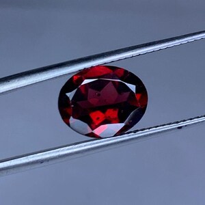 May include: A faceted, oval-shaped, deep red gemstone. The gem is held by a pair of silver tweezers against a blurred gray background. The gem's facets reflect light, creating a sparkling effect. This gemstone could be used in jewelry.
