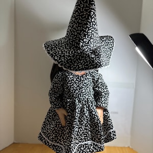 May include: A doll dressed in a black and white witch costume. The dress and tall hat are patterned with white bones on a black background. The dress has long sleeves and a white lace trim. The doll wears white shoes.