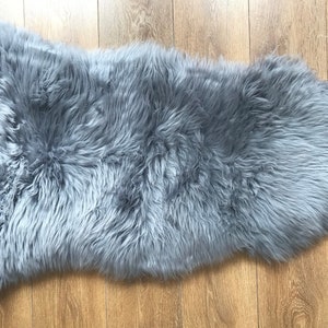 May include: A soft, gray sheepskin rug with long, fluffy fur. The rug is laid out flat on a wooden floor.