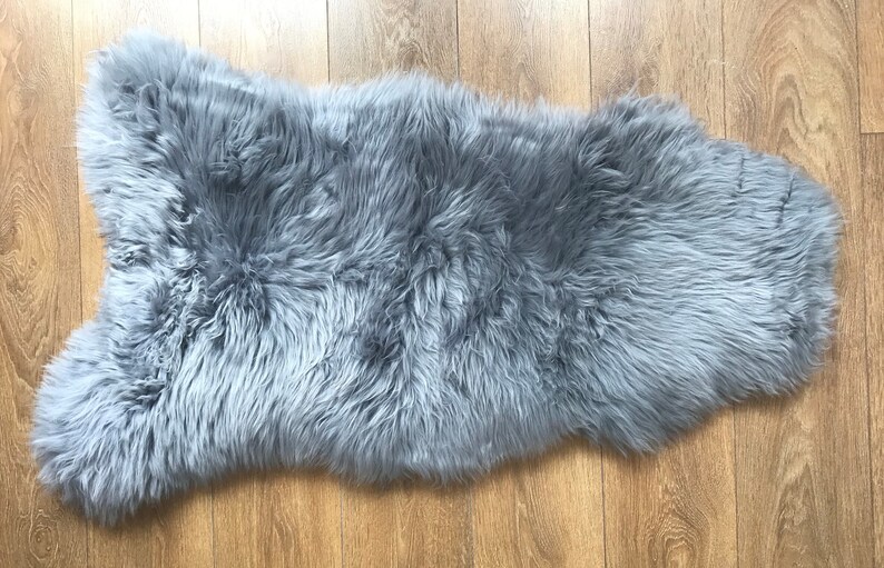 May include: A soft, gray sheepskin rug with long, fluffy fur. The rug is laid out flat on a wooden floor.