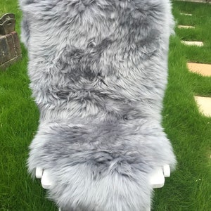 May include: A soft, gray sheepskin rug with long, fluffy fur. The rug is draped over a white folding chair and is sitting on a green grassy lawn.