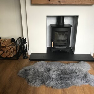 May include: A black wood-burning stove with a glass door and a wooden handle is set into a white fireplace with a black slate hearth. A gray sheepskin rug is in front of the fireplace. A black metal log holder with logs is on the left side of the image.