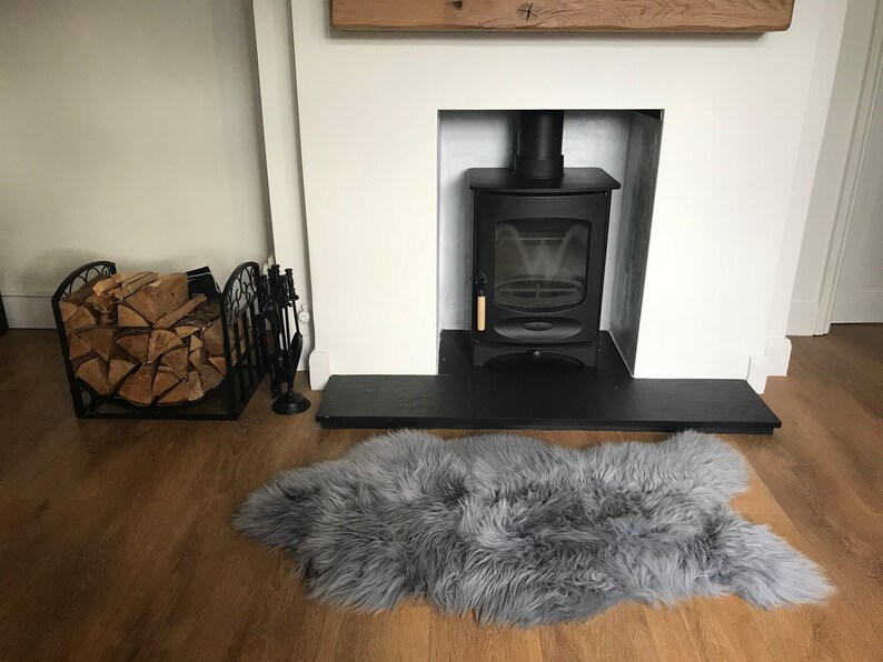 May include: A black wood-burning stove with a glass door and a wooden handle is set into a white fireplace with a black slate hearth. A gray sheepskin rug is in front of the fireplace. A black metal log holder with logs is on the left side of the image.