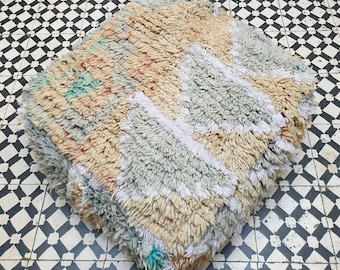 May include: A fluffy, hand-woven pouf with a geometric pattern in shades of beige, light blue, and white. The pouf is made of wool and is perfect for adding a touch of Moroccan style to your home.