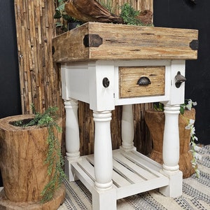 May include: A rustic butcher block table with a white base and a natural wood top. The table features a drawer, a bottle opener, and a lower shelf. Decorative elements include greenery and a wooden log.
