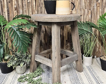 May include: A rustic wooden stool with a round top and three legs. The stool is weathered and has a natural wood finish. It is sitting on a white and black patterned rug. There are potted plants and a mug on the stool.