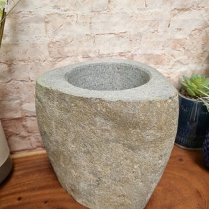 May include: A gray stone vase with a wide, oval opening. The vase has a rough, textured surface and a natural, earthy appearance. The vase is on a wooden surface.