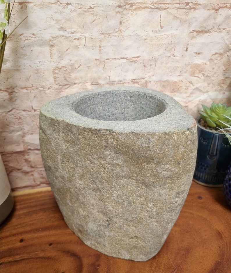 May include: A gray stone vase with a wide, oval opening. The vase has a rough, textured surface and a natural, earthy appearance. The vase is on a wooden surface.